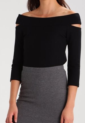 Femme portant un haut noir épaules dénudées avec des manches trois-quarts et une jupe crayon grise taille haute.