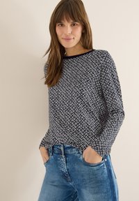 Granatowy top z długim rękawem z geometrycznym białym wzorem. Okrągły dekolt i luźny krój, zestawiony z jasnoniebieskimi jeansami z przednimi kieszeniami.