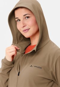 Beige Zip-Hoodie aus strukturiertem Stoff, mit Kapuze und dem "VAUDE"-Logo auf der Brust. Ein orangenes Shirt ist darunter sichtbar.
