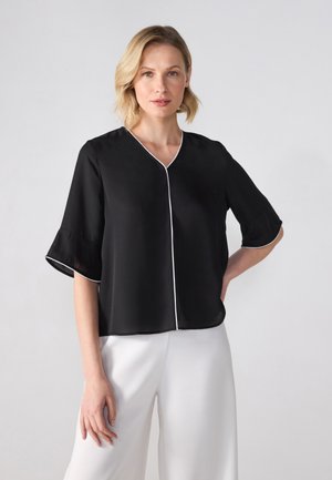 Blusa nera a maniche corte con scollatura a V e piping bianco lungo i bordi. Tessuto liscio con una vestibilità ampia, abbinata a pantaloni bianchi.
