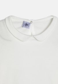 Weißes Baumwolloberteil mit rundem Kragen und Knopfverschluss auf der Rückseite; zeichnet sich durch eine glatte Textur und ein minimalistisches Design aus. Pflegeetikett sichtbar.