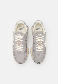 Pelēki zamšādas sporta apavi ar austām tekstūrām, baltām šņorēm, bēšiem akcentiem un melnu gumijas zoli, ar New Balance logo uz mēles.