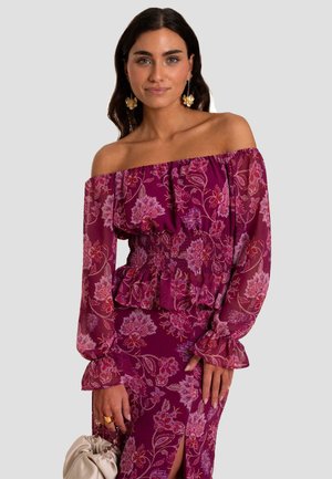 Robe décolletée en violet profond avec des motifs floraux, taille smockée, manches volantes et fente sur le côté. Tissu lisse et fluide.