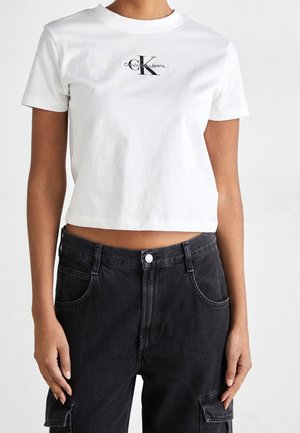 Kobieta ubrana w białą przyciętą koszulkę Calvin Klein Jeans i czarne, wysokowysoko taliowane spodnie cargo z przednimi kieszeniami.