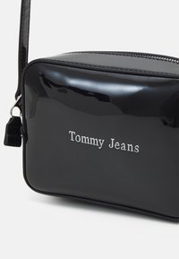 Borsa rettangolare in pelle nera lucida con chiusura a zip. Presenta il logo argentato "Tommy Jeans" sulla parte frontale e una tracolla regolabile.
