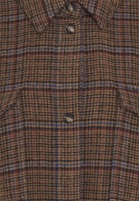 Bruin geruit wollen shirt met blauwe en rode lijnen, voorzien van donkere knopen en twee voorkruiszakken onder de kraag.