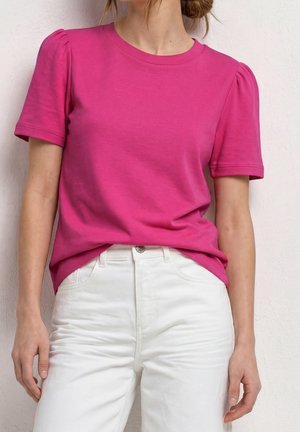 Donna che indossa una camicia rosa acceso a maniche corte con spalle imbottite e pantaloni bianchi a vita alta, in piedi contro una parete chiara.