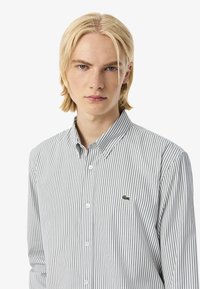 Giovane con capelli lunghi e lisci biondi, che indossa una camicia a righe con un piccolo logo. Espressione seria, sfondo bianco.