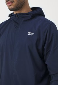 Tumši zilas krāsas vieglais džemperis ar rāvējslēdzēju priekšā, regulējamu kapuci un diskrētu Reebok logo uz krūtīm. Izgatavots no gludas, elpojošas materiāla.