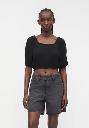 Femeie cu păr împletit purtând un top scurt negru cu mâneci bufante și pantaloni scurți din denim negru șters, cu talie înaltă, stând în picioare pe un fundal simplu.