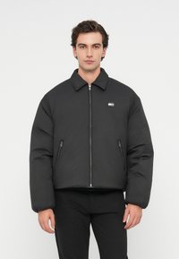 Mustapaitainen toppatakki, jossa on kaulus, etuvetoketju, sivuvetoketjulliset taskut ja logoyksityiskohta rintaketjussa. Sileä pinta, lyhennetty malli.