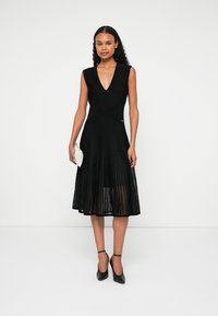 Μαύρο πλισέ φόρεμα με βαθύ V-ντεκολτέ, χωρίς μανίκια και εφαρμοστή μέση, συνδυασμένο με μια μικρή ανοιχτόχρωμη τσάντα και μαύρα τακούνια.