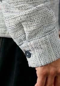 Lys blå tekstureret stof med et mønstret væv og en knaplukning ved manchetten, der fremhæver det detaljerede håndværk og det bløde udseende.