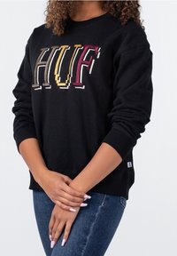 Felpa nera con ricamo colorato "HUF" in diverse stili di carattere. Il tessuto è morbido, con polsini a coste e una vestibilità rilassata.