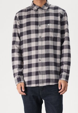 Mężczyzna ubrany w czarno-szarą kraciastą koszulę z długim rękawem zapinaną na guziki z kieszenią na piersi oraz ciemne spodnie, stojący na jednolitym tle.