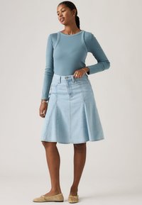 Γυναίκα που φοράει μπλούζα με μακριά μανίκια σε πετρόλ απόχρωση, ανοιχτό μπλε τζιν φούστα μήκους μέχρι το γόνατο, χρυσές μπαλαρίνες, με πλεγμένα μαλλιά και λιτά κοσμήματα, στέκεται μπροστά σε ουδέτερο φόντο.