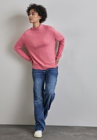 Pull en tricot rose avec un col montant, associé à un jean évasé bleu clair. La tenue présente des poignets côtelés et une coupe décontractée. Des baskets blanches viennent compléter le look.