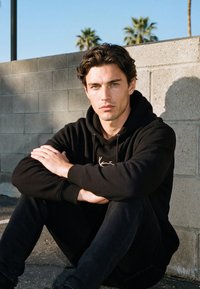 Giovane uomo con felpa nera con cappuccio e pantaloni, seduto a gambe incrociate contro un muro di cemento all'aperto con palme sullo sfondo.