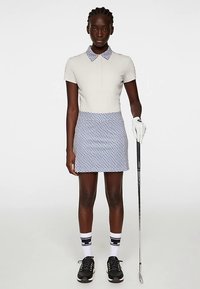 Golfantrekk med en tettsittende beige poloskjorte, et blåmønstret skjørt, svarte joggesko, knelange sokker og en golfkølle.