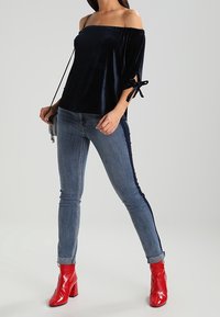 Marinblå off-shoulder sammetstopp med bandknutar, matchat med ljusblå smala jeans med en mörk sidostråk, och klarröda klackstövlar.