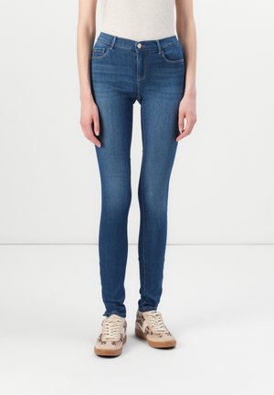 Persoon draagt blauwe strakke jeans en beige sneakers met luipaardprint, staand tegen een effen witte achtergrond.