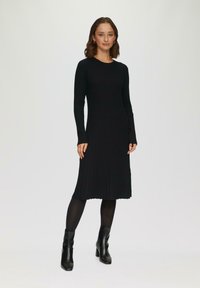 Zwarte ribgebreide jurk met lange mouwen en een ronde halslijn, tot halverwege de kuit. Draagt met zwarte enkellaarsjes en panty’s. Eenvoudig ontwerp.