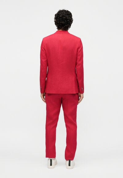 Άτομο με σγουρά μαλλιά στέκεται γυρισμένο με την πλάτη του, φοράει κόκκινο κοστούμι και λευκά αθλητικά παπούτσια με μαύρες ρίγες στις φτέρνες, σε λευκό φόντο.