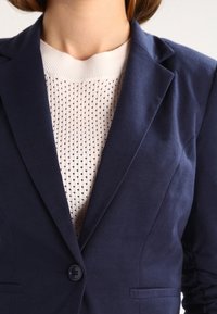 Námořnický modrý blazer s texturovanou tkaninou, s jedním knoflíkem a klopami. Pod ním je smetanový pletený top s malými perforacemi.