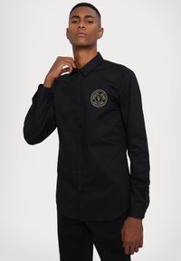 Camicia nera con bottoni e maniche lunghe, caratterizzata da un logo circolare dorato sul lato sinistro del petto e una texture di tessuto liscia e uniforme.