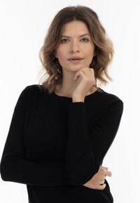 Pulover negru tricotat cu guler rotund și măneci lungi. Modelul stă în poziție cu mâna pe bărbie, pe un fundal alb simplu.