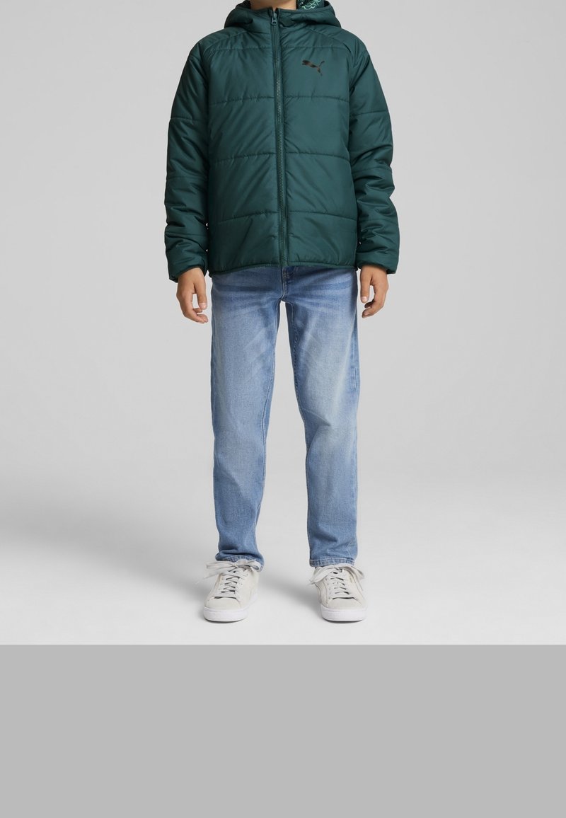 Enfant portant une doudoune à capuche vert foncé, un jean bleu clair et des baskets blanches, debout devant un fond gris uni.