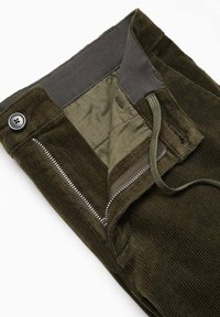 Pantalon en velours côtelé vert foncé avec braguette zippée ouverte, fermeture par bouton et doublure intérieure visible.