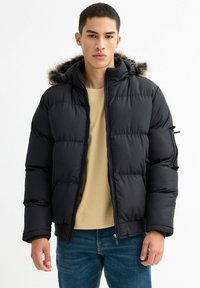 Chaqueta de plumas negra con capucha de piel sintética, diseño acolchado y bolsillos laterales con cremallera, combinada con una camisa beige y jeans azules.