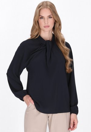 Blusa blu navy realizzata in tessuto leggero con colletto a fiocco, maniche lunghe con polsini arricciati e vestibilità rilassata. Abbinata a pantaloni beige.