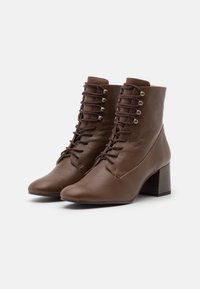 Par de botas de cuero marrón hasta el tobillo con tacones de bloque y diseño de cordones en la parte delantera sobre un fondo blanco.