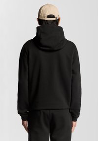 Felpa nera con cappuccio e texture morbida, caratterizzata da un ampio cappuccio e polsini a costine. Indossata con pantaloni neri abbinati, vista da dietro.