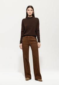 Brauner Strickpullover mit hohem Kragen, kombiniert mit braunen weiten Hosen, die goldene Knopfakzente an den Taschen haben.