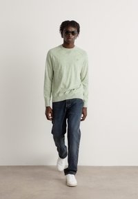 Sweatshirt lung cu mânecă lungă, de culoare verde deschis, cu manșete cu nervuri, purtat împreună cu blugi negri și adidași albi. Modelul poartă ochelari de soare închiși la culoare, stând pe un fundal simplu.