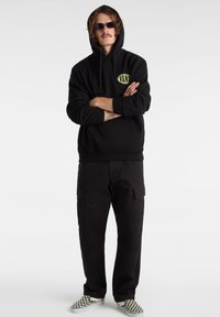 Felpa nera con cappuccio e logo giallo Vans, abbinata a pantaloni cargo neri e scarpe slip-on a scacchi bianco e nero.
