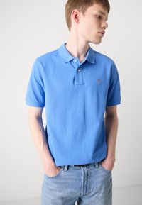 Helles blaues Poloshirt aus Baumwollstoff, mit klassischem Kragen, drei Knöpfen und kleinem gesticktem Logo auf der Brust. Dazu werden Jeans aus Denim getragen.