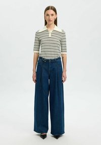 Randig kortärmad polo i krämfärgad och svart; vida mörk denimbyxor med knytbälte; modell bär spetsiga svarta klackar.