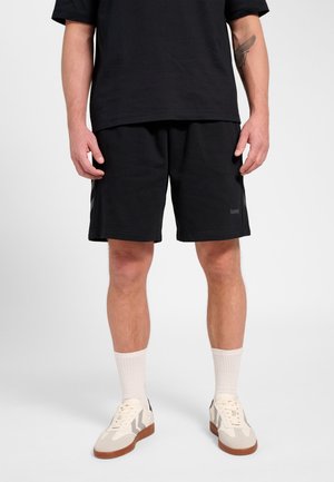 Mand iført sorte shorts, sort t-shirt, hvide ankel strømper og beige sneakers, står op mod en ensfarvet baggrund.