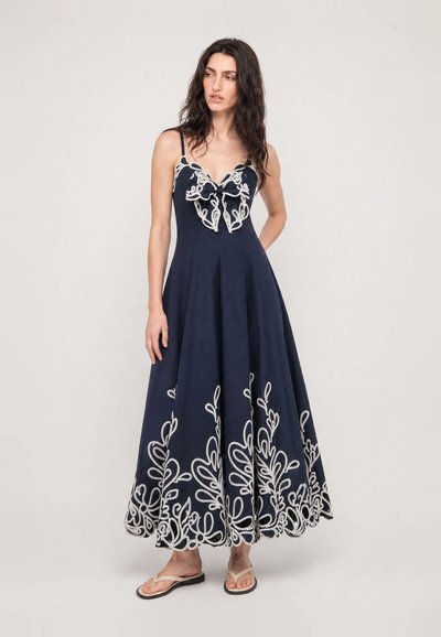 Femme portant une robe longue sans manches bleu marine avec broderie florale blanche et des tongs beiges, debout devant un fond uni.