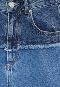 Gros plan de deux tissus en denim bleu cousus ensemble, avec bouton visible, poche, passant de ceinture et bord brut effiloché entre les tissus.