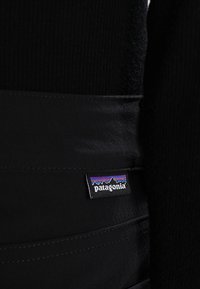 Section de tissu noir avec une petite étiquette rectangulaire de la marque Patagonia présentant un logo de montagne en violet, bleu, rouge et blanc.