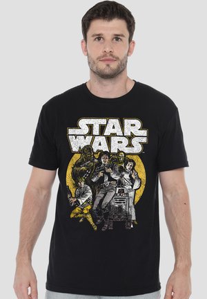Mann trägt schwarzes T-Shirt mit Vintage Star Wars Charakteren und Logo in Weiß und Gelb vorne, vor einfarbig grauem Hintergrund.