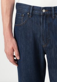 Jeans de mezclilla azul oscuro con corte recto, que presentan un diseño de cinco bolsillos, costuras en contraste y un cierre de botón metálico en la cintura.