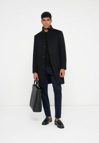 Hombre vestido con un abrigo negro de botones, pantalones marinos, mocasines negros con borlas, sosteniendo un bolso tote de cuero negro, de pie frente a un fondo blanco.