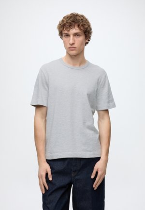 Jeune homme aux cheveux bouclés portant un t-shirt uni gris clair à manches courtes et un jean bleu foncé, debout devant un fond blanc.
