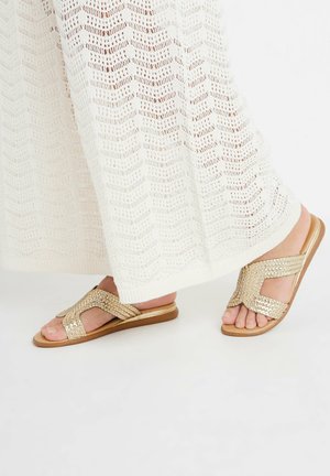 Sandales en fil d'or avec un design à bout ouvert et à lanières croisées, posées sur une semelle plate, accompagnées d'un tissu blanc texturé.